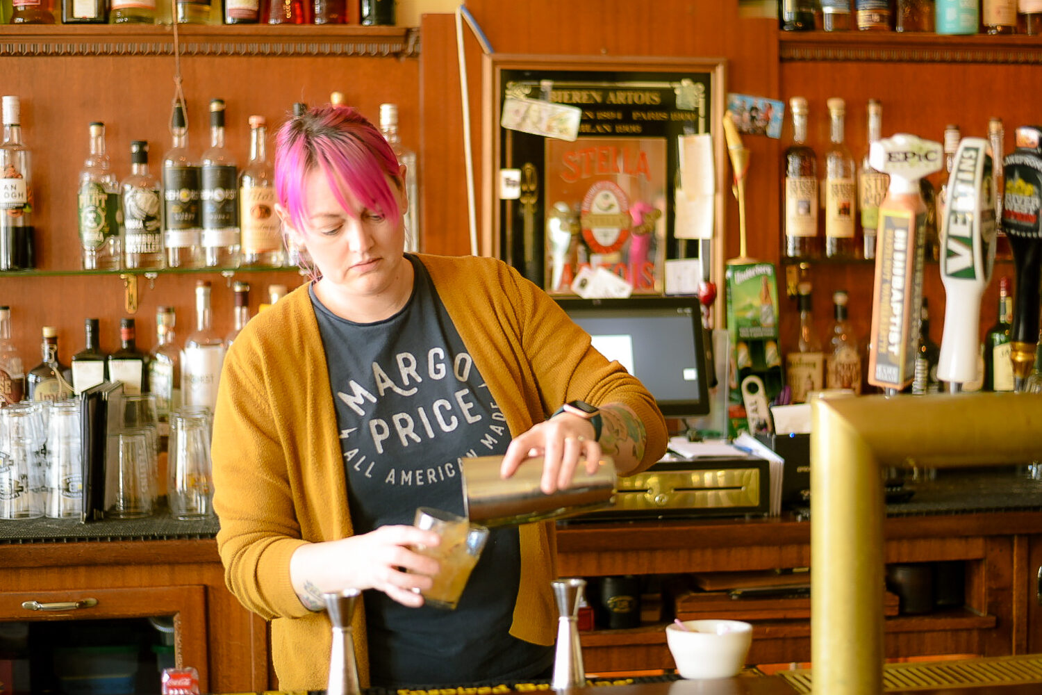woman with pink hair behind a bar pouring a drink from a metal shaker into a tumbler glass