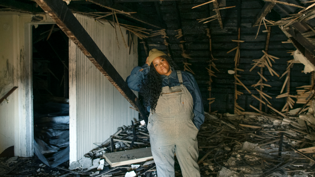 Artist portrait of vanessa german. german is standing inside a building that's in extreme disrepair, looking at the camera.