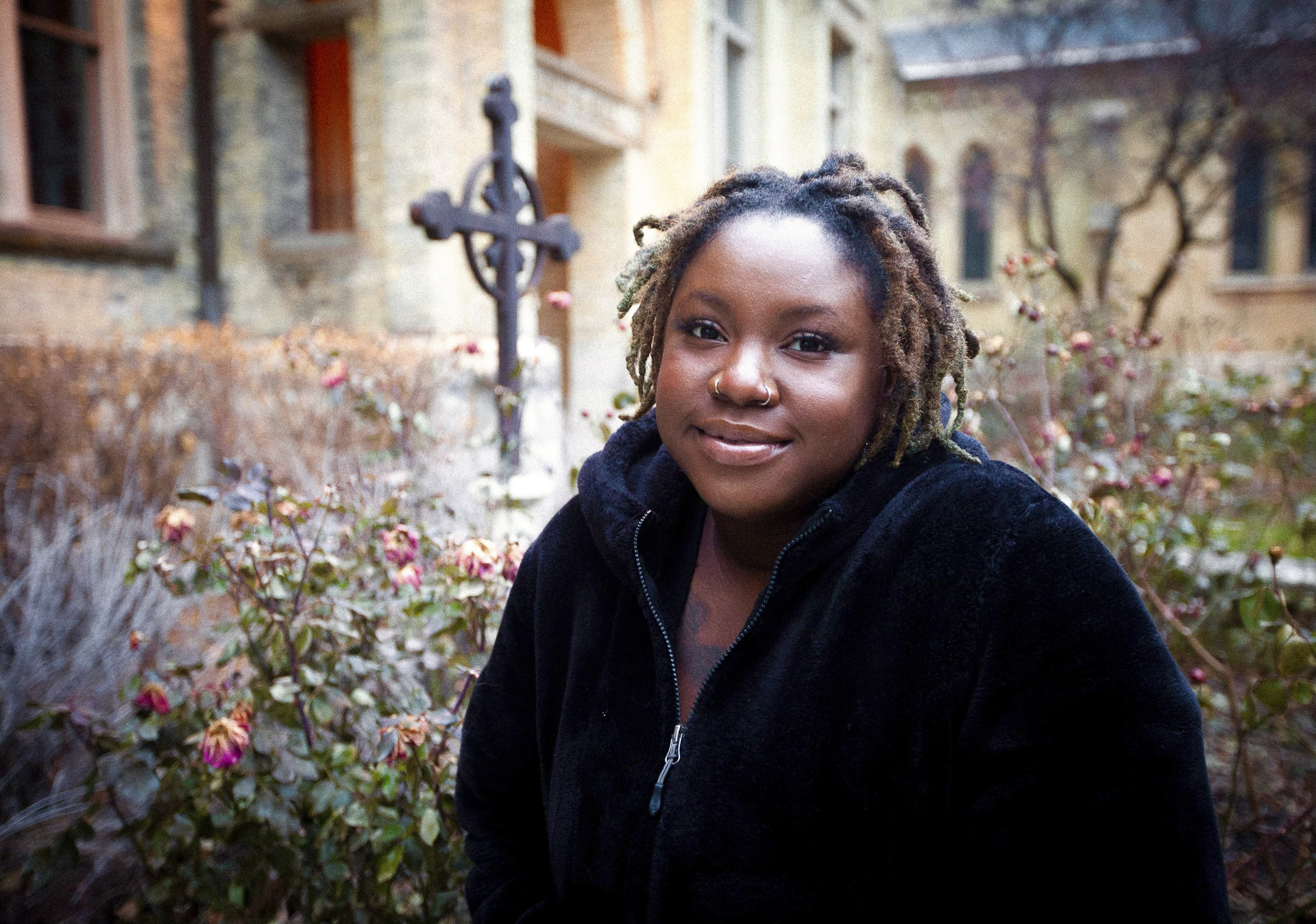 Ladasia Bryant artist portrait. Ladasia photographed outside, wildflowers, a cross, and a stone building are in the background.